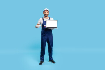 Full length expert worker man in uniform pointing at laptop with mock up blank display, showing empty place for advertise, message or repair service order. studio shot isolated on blue background