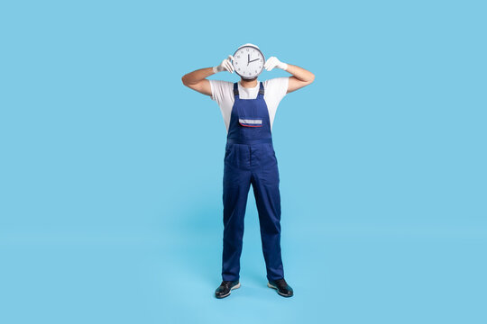 Full length anonymous labourer in uniform covering face with clock. Advertisement of professional house repair services on time, punctual courier delivery. studio shot isolated on blue background