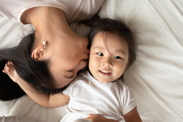 Above top view affectionate asian ethnicity mother kissing little cute pretty biracial kid daughter, relaxing on comfortable bed, enjoying tender morning time together at home, head shot close up.