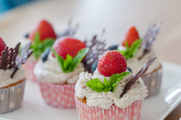 Selected focus view at beautiful and colourful group of cupcakes decorated with white whip cream, strawberries, blueberries, chocolate and mint on wooden table. 
