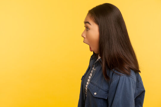 Oh my god, wow! Side view of excited girl in denim shirt standing with mouth open in surprise, shouting in amazement, shocked by unbelievable success. indoor studio shot isolated on yellow background