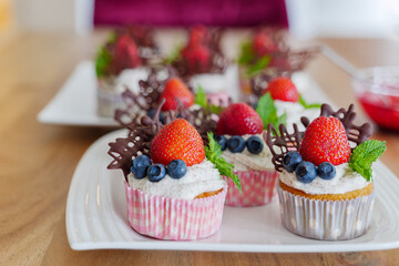 Selected focus view at beautiful and colourful group of cupcakes decorated with white whip cream, strawberries, blueberries, chocolate and mint on wooden table. 