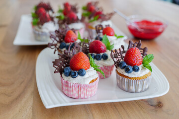 Selected focus view at beautiful and colourful group of cupcakes decorated with white whip cream, strawberries, blueberries, chocolate and mint on wooden table. 