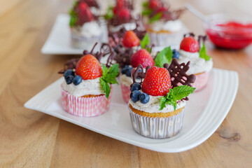 Selected focus view at beautiful and colourful group of cupcakes decorated with white whip cream, strawberries, blueberries, chocolate and mint on wooden table. 