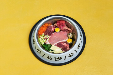 Bowl of natural organic raw food for dog on yellow background Meat, chicken head, vegetables, quail egg and butter