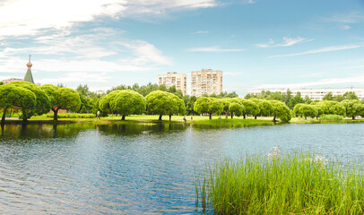 Obraz premium Pond in a city park with a series of lush willows on the shore and a church and high-rise buildings in the background.