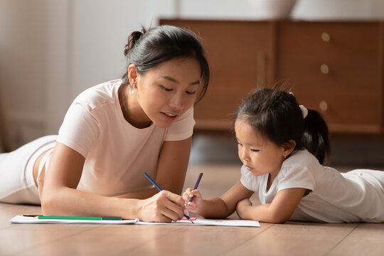 Affectionate caring vietnamese ethnicity woman lying on floor with cute small preschool biracial child daughter, involved in hand drawing pictures together in paper album, childcare hobby pastime.