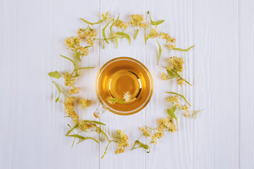 Top view on fresh herbal linden tea in transparent glass cup with dry linden flowers around on white wooden background with copyspace. Organic herbal infusion in alternative folk medicine concept