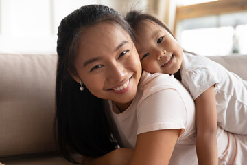 Head shot portrait smiling attractive young asian mother resting on sofa, holding on back cute small preschool vietnamese ethnicity child daughter, enjoying relaxed free time, looking at camera.