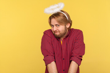 Funny modest cute bearded guy with saint nimbus over head looking at camera with shy timid...
