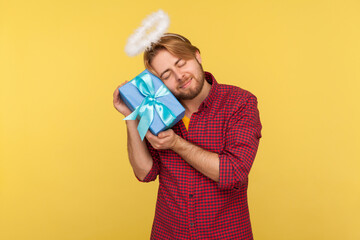 Funny cute angelic bearded guy with saint halo on head embracing gift box, admiring perfect holiday...