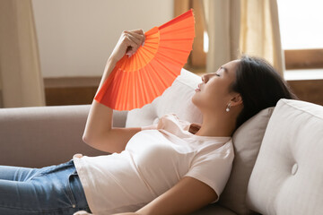 Side view tired young asian ethnicity woman lying on sofa, using paper fan indoors, unhappy...