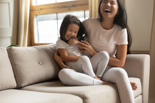 Overjoyed young vietnamese ethnic woman mother sitting on cozy couch, tickling little adorable funny preschool asian kid daughter, enjoying playtime together at home, laughing joking indoors.