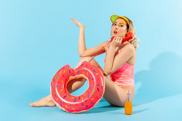 Full length, indignant angry woman in bathing suit sitting with rubber ring, cocktail and talking on phone handset with resentful annoyed expression. indoor studio shot isolated on blue background