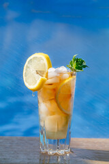 A glass of ice tea with lemon and mint, poolside with blue water in the background
