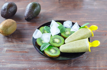 Home made healthy sorbet (fruit ice cream) made of avocado and kiwi on the black plate among pieces of kiwi, mint leaves, ice cubes on the old rustic wooden table near whole avocados and kiwi - obrazy, fototapety, plakaty