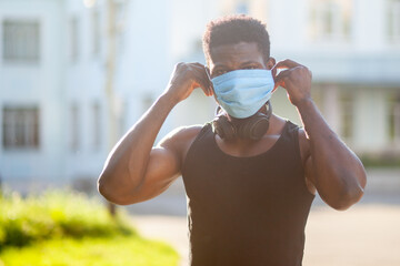 African American guy puts on a medical mask outdoors against the background of the city, people in the coronavirus pandemic - Powered by Adobe