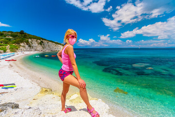 Blonde woman with pink dress and surgical mask on promontory looking Padulella beach near Portoferraio during Covid-19. Tourist in holidays on Elba island, Tuscany. Travel in Italy with Coronavirus.
