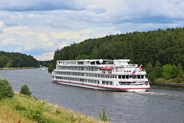 Russian four deck white passenger ship, cruise liner floating on Moscow river Canal, tourist vessel rear side view on summer day with green trees on rivershore and blue cloudy sky background
