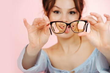 Eccentric young woman holding glasses in front of her face, sticking tongue out. Image triples in lenses. Over pink background.