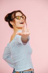 Cute eccentric provocative girl looking at camera, inviting with a finger. Over pink background. She wears glasses, two buns on her head and a shirt with an open top. Focus on a hand.