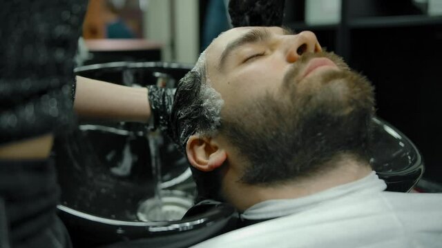 Closeup of a head wash in a barber shop with gloves on. A young bearded man with his eyes closed is waiting for a haircut. Handsome girl hairdresser washing his head. Slow motion