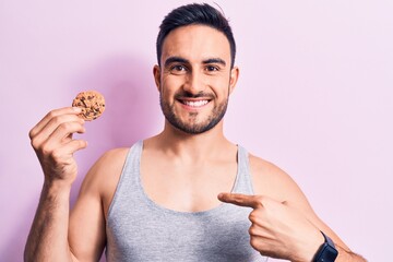 Young handsome man with beard wearing sleeveless t-shirt eating chocolate cookie pointing finger to one self smiling happy and proud