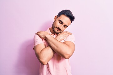 Young handsome man with beard wearing casual polo standing over pink background hugging oneself happy and positive, smiling confident. Self love and self care