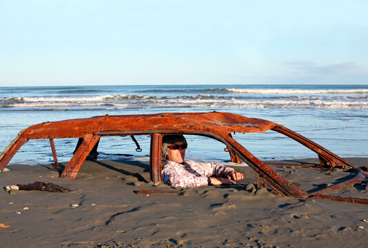 Man sits in rusted car wreck buried in sand on beach as the tide comes in