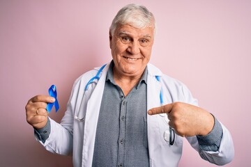 Senior handsome hoary doctor man wearing stethoscope holding blue cancer ribbon with surprise face pointing finger to himself