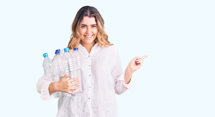 Obraz premium Young caucasian woman holding recycling plastic bottles smiling happy pointing with hand and finger to the side