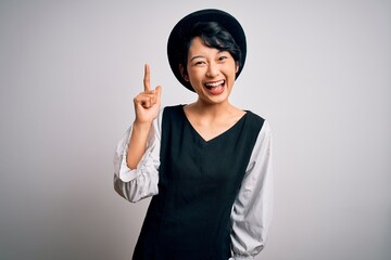 Young beautiful asian girl wearing casual dress and hat standing over isolated white background pointing finger up with successful idea. Exited and happy. Number one.