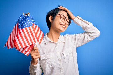 Young beautiful asian patriotic girl holding united states flags celebrating independence day stressed with hand on head, shocked with shame and surprise face, angry and frustrated. Fear and upset.