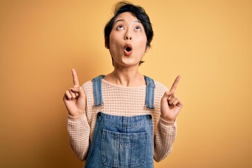 Young beautiful asian girl wearing casual denim overalls over isolated yellow background amazed and surprised looking up and pointing with fingers and raised arms.