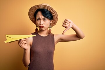 Young beautiful chinese woman on vacation wearing summer hat holding paper airplane with angry face, negative sign showing dislike with thumbs down, rejection concept