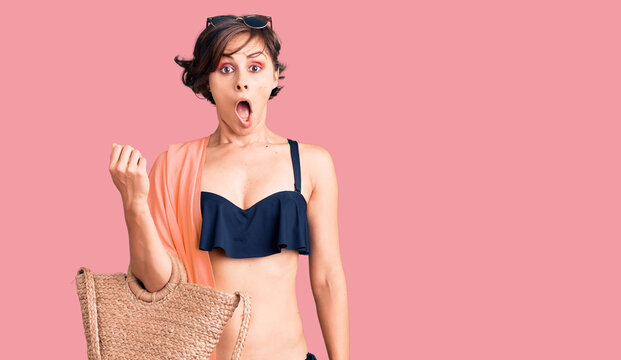 Beautiful young woman with short hair wearing bikini and hat holding summer wicker handbag scared and amazed with open mouth for surprise, disbelief face