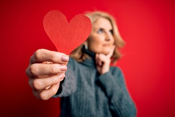 Middle age beautiful blonde romantic woman holding red paper heart celebrating valentine day serious face thinking about question, very confused idea