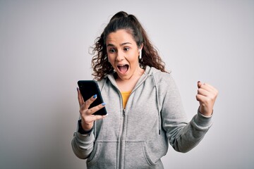 Beautiful sportswoman with curly hair listening to music using smartphone and earphones screaming proud and celebrating victory and success very excited, cheering emotion