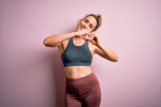Young beautiful blonde sportswoman doing sport wearing sportswear over pink background smiling in love doing heart symbol shape with hands. Romantic concept. © Krakenimages.com