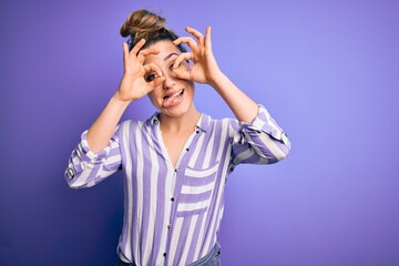 Young beautiful blonde woman wearing casual striped shirt standing over purple background doing ok gesture like binoculars sticking tongue out, eyes looking through fingers. Crazy expression.