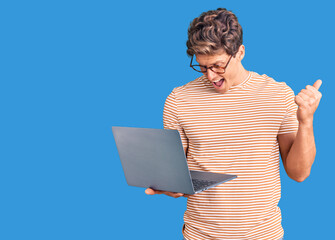 Young handsome man wearing glasses holding laptop screaming proud, celebrating victory and success very excited with raised arms - obrazy, fototapety, plakaty