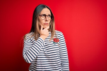 Young beautiful blonde woman with blue eyes wearing glasses standing over red background Thinking worried about a question, concerned and nervous with hand on chin