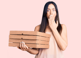 Young beautiful caucasian woman holding delivery pizza box covering mouth with hand, shocked and...