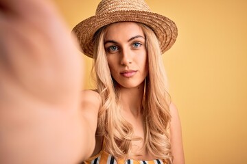 Young beautiful blonde woman on vacation wearing bikini and hat making selfie by camera with a confident expression on smart face thinking serious