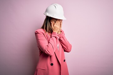 Young beautiful brunette architect woman wearing safety helmet over pink background with sad expression covering face with hands while crying. Depression concept.