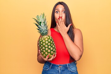 Young beautiful brunette woman holding pineapple covering mouth with hand, shocked and afraid for...