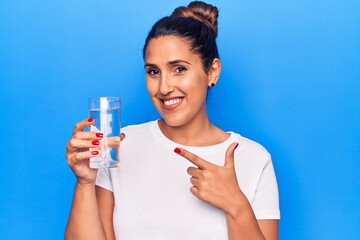 Young beautiful brunette woman drinking glass of water smiling happy pointing with hand and finger