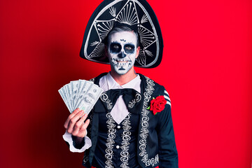 Young man wearing mexican day of the dead costume holding dollars looking positive and happy standing and smiling with a confident smile showing teeth