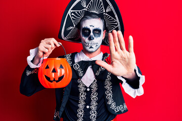 Young man wearing mexican day of the dead costume holding pumpkin with open hand doing stop sign with serious and confident expression, defense gesture
