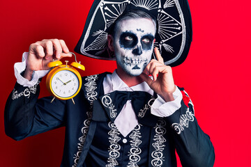 Young man wearing mexican day of the dead costume holding alarm clock serious face thinking about question with hand on chin, thoughtful about confusing idea